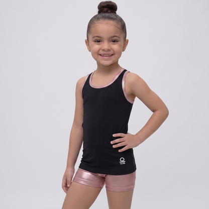 A young girl with her hair in a high bun stands against a light gray studio background, wearing a black sleeveless athletic tank top with metallic pink trim and matching metallic pink shorts. A small white logo is visible near the lower left hem of the tank top.