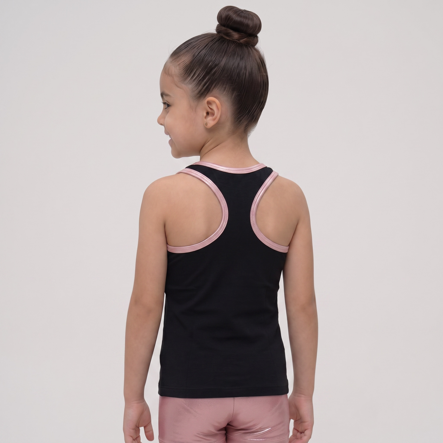 A young girl with her hair in a high bun is shown from the back, wearing a black athletic tank top with light pink trim around the shoulder straps and neckline, paired with matching pink athletic shorts, against a light gray studio background.