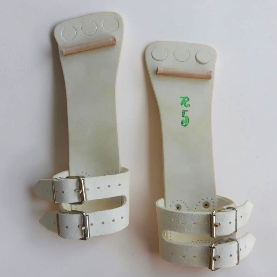 A pair of off-white leather wrist wraps or splints with silver metal buckles, displayed against a plain light background. One has green printed characters, and both feature circular cutouts near the top.