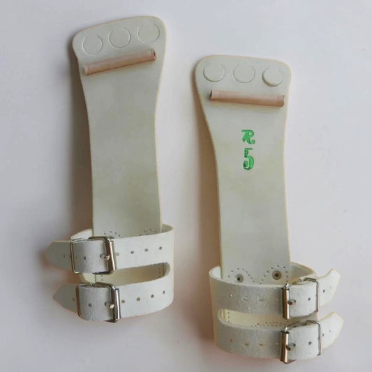 A pair of off-white leather wrist wraps or splints with silver metal buckles, displayed against a plain light background. One has green printed characters, and both feature circular cutouts near the top.